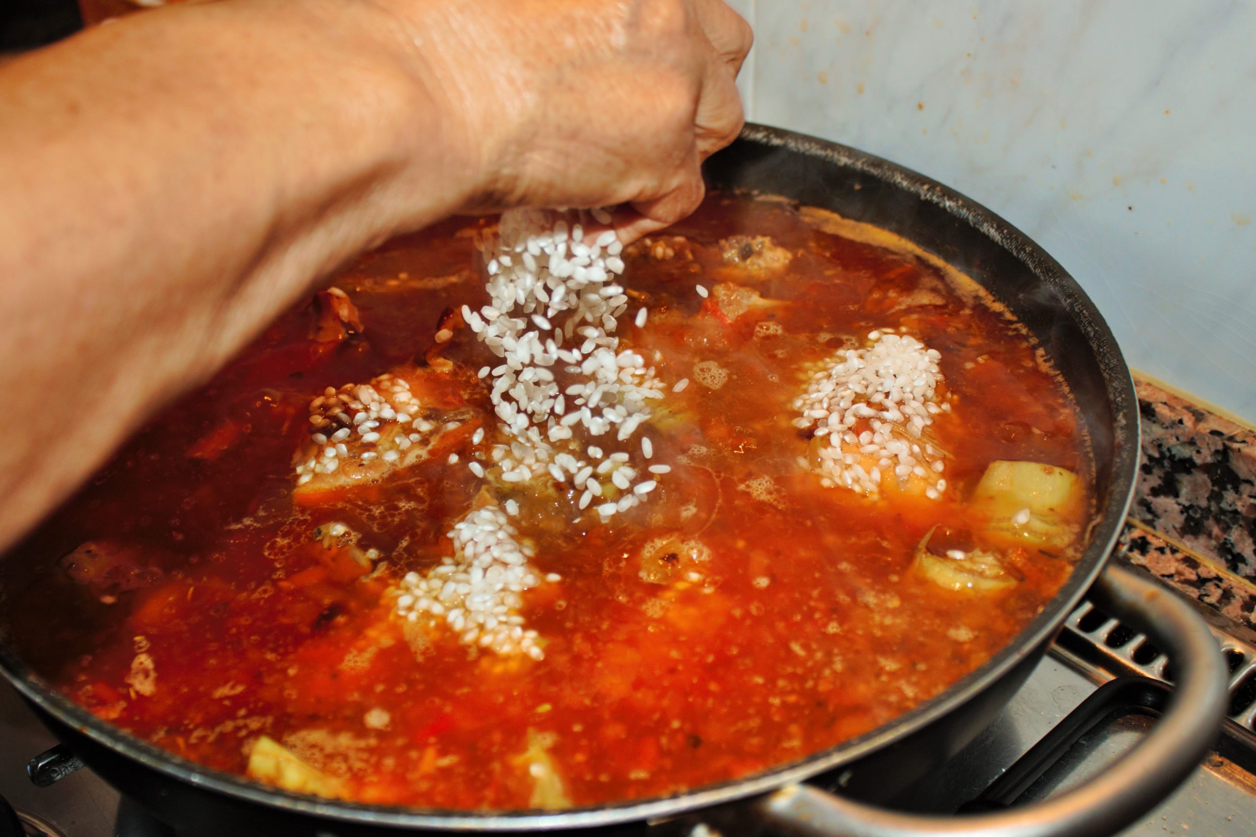 Añadiendo el arroz a la paella para que se impregne del sofrito de conejo y alcachofas