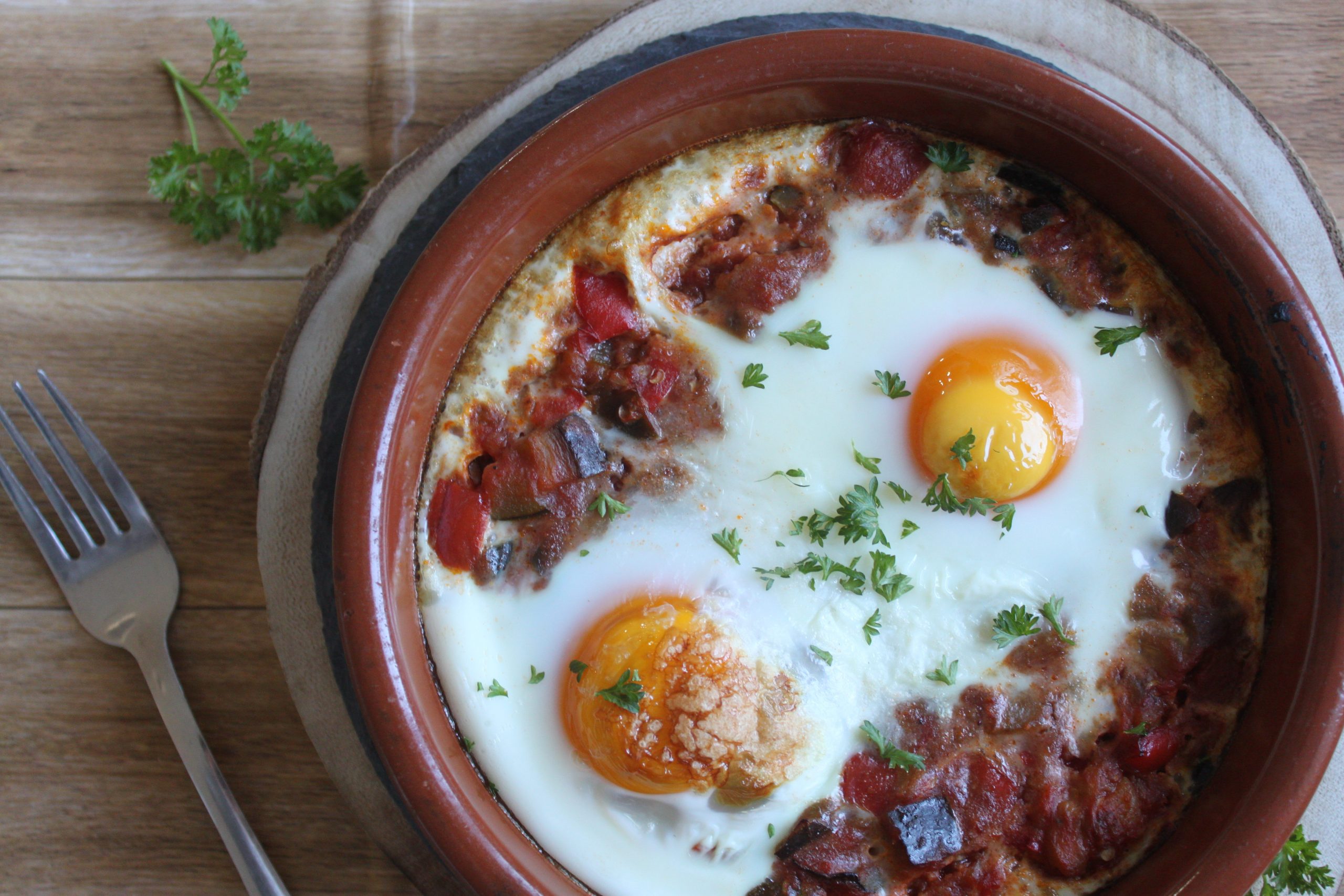 Cazuela de barro con huevos al horno sobre una cama de sanfaina de verduras brillante y perejil fresco.