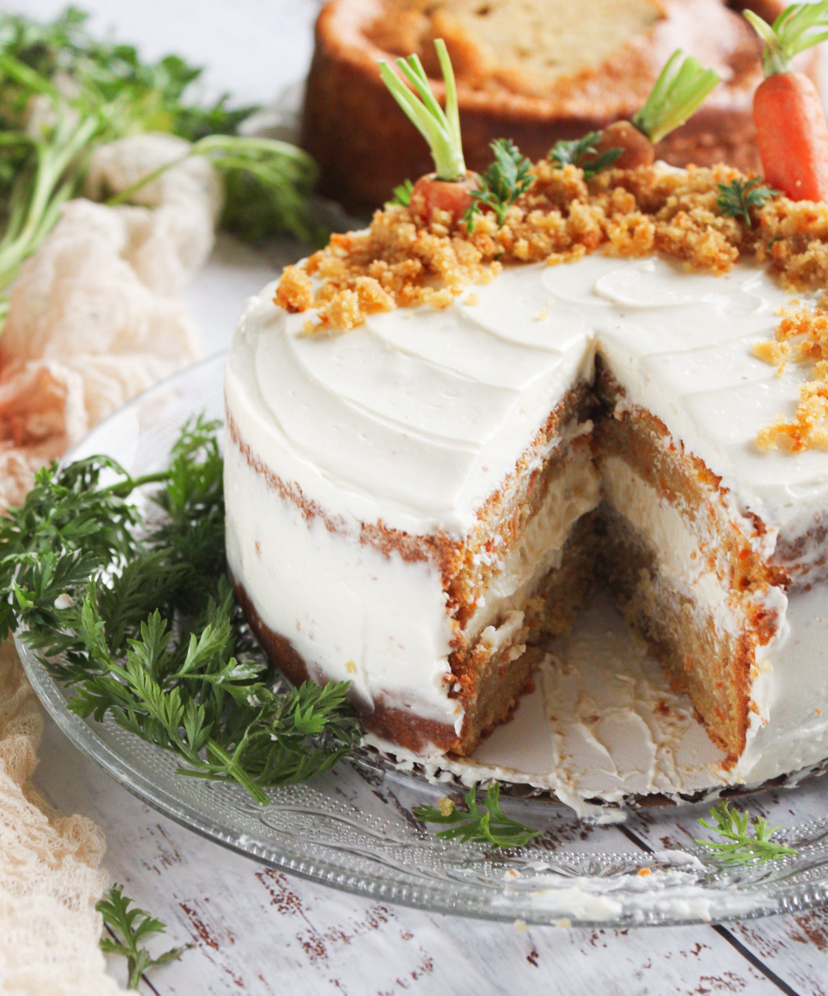 Tarta de zanahoria r&uacute;stica con cobertura de crema de queso, donde se ve el interior de la tarta y est&aacute; decorada con zanahorias baby frescas y hojas verdes sobre una mesa de madera blanca r&uacute;stica