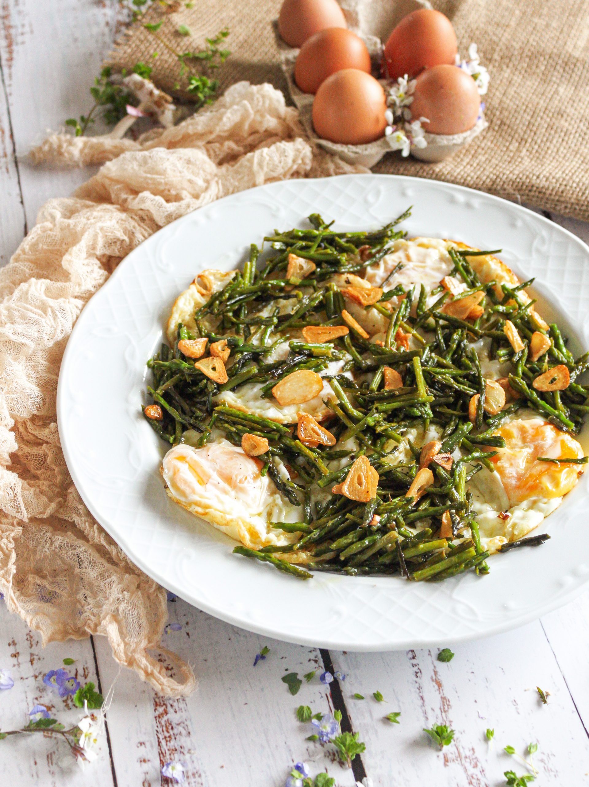 Primer plano de unos huevos fritos con puntilla dorada y yema líquida, servidos sobre una base de espárragos trigueros salteados con láminas de ajo crujiente.