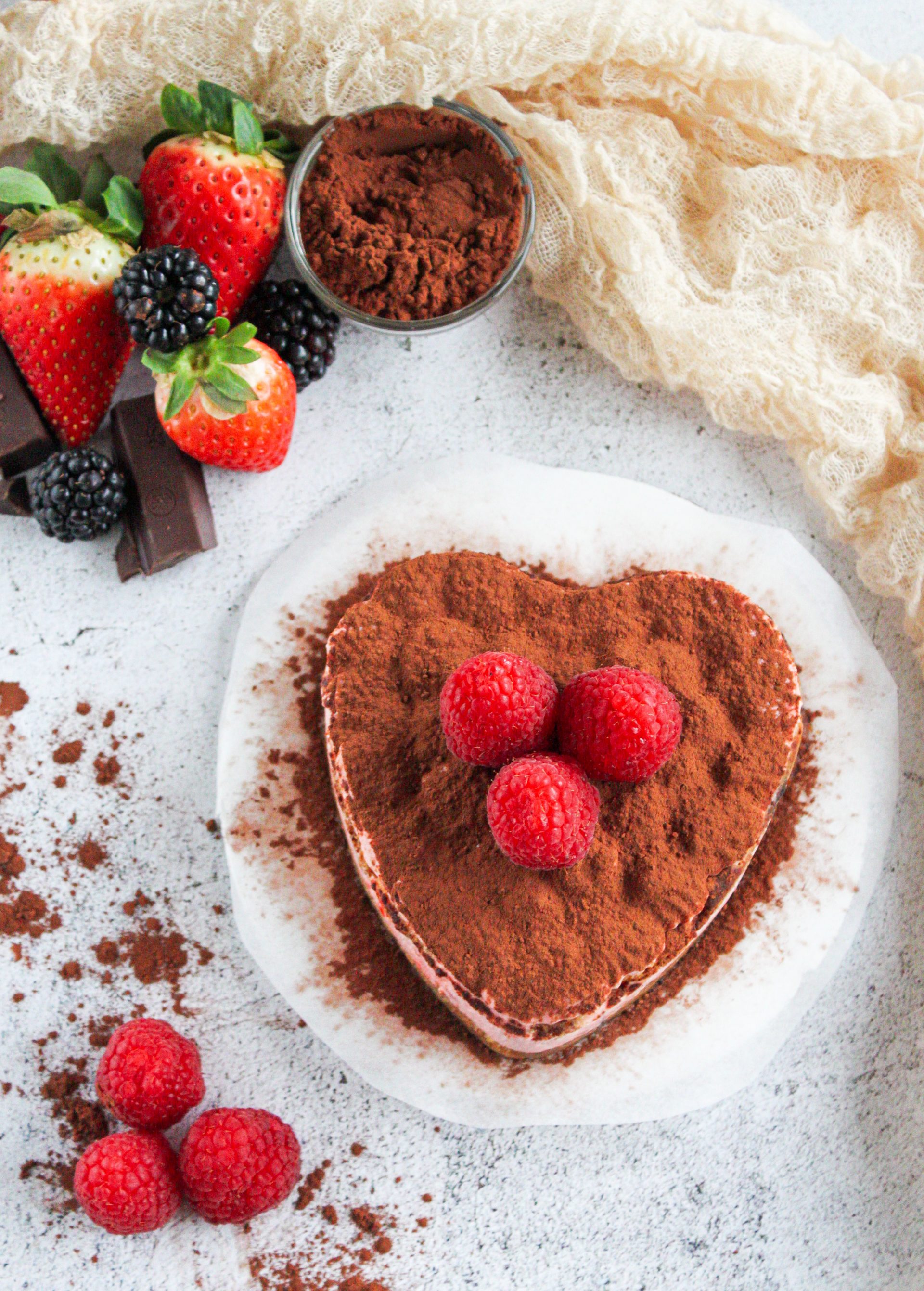 Corazón de tiramisú decorado con frambuesas y cacao en polvo, servido con frutas frescas de decoración.