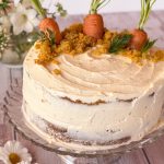 Tarta de zanahoria casera entera decorada con ramas de zanahoria en la base, sobre un plato de cristal y fondo rústico de madera blanca.
