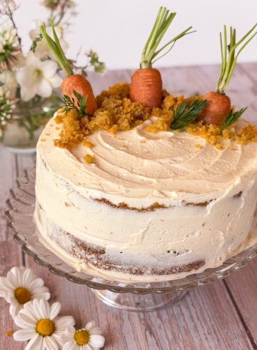 Tarta de zanahoria casera entera decorada con ramas de zanahoria en la base, sobre un plato de cristal y fondo rústico de madera blanca.