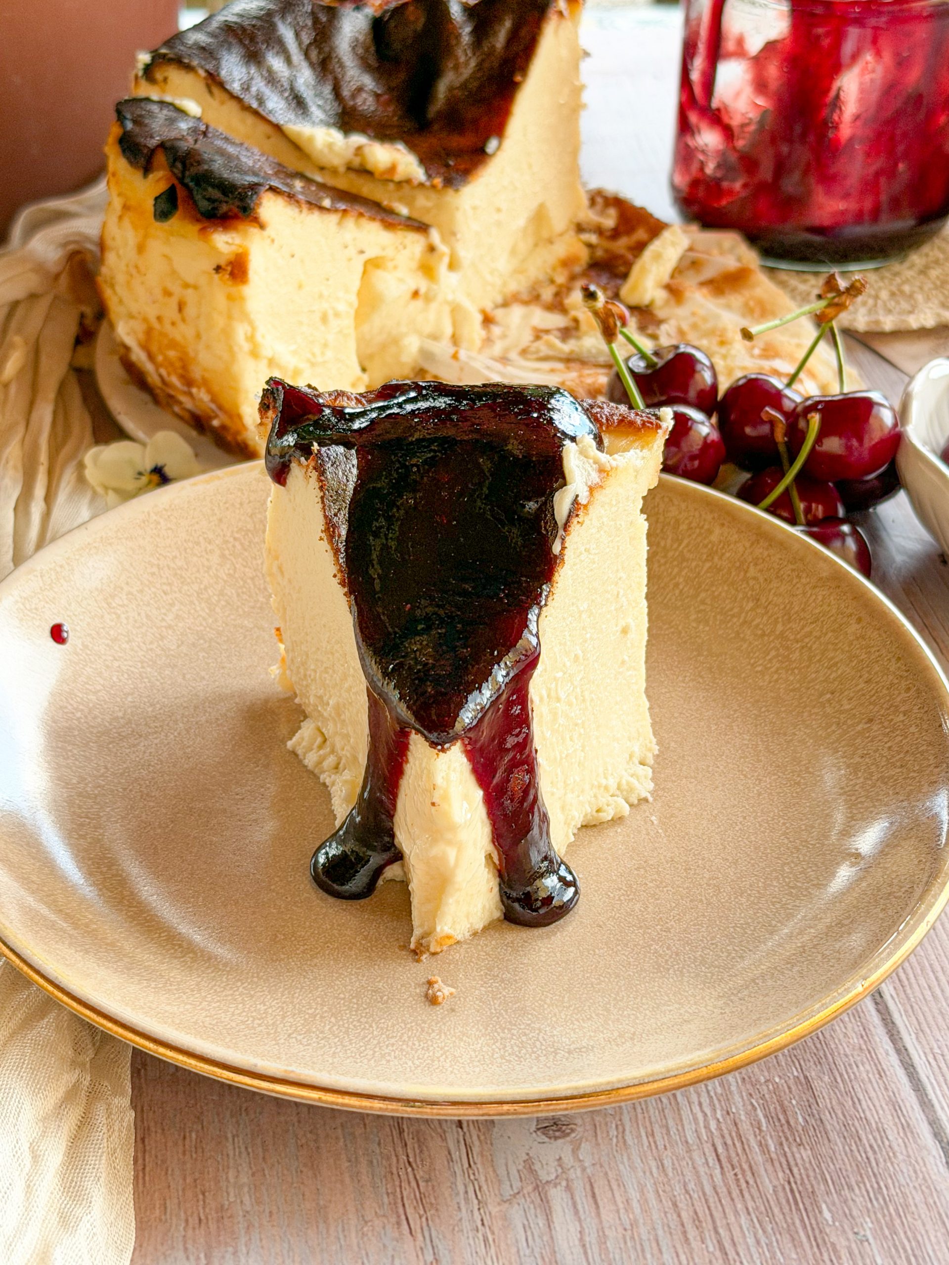 Primer plano cenital de una tarta de queso estilo La Viña con el centro fundente, mientras se vierte una mermelada casera de cerezas brillantes y trozos enteros de fruta sobre su superficie tostada