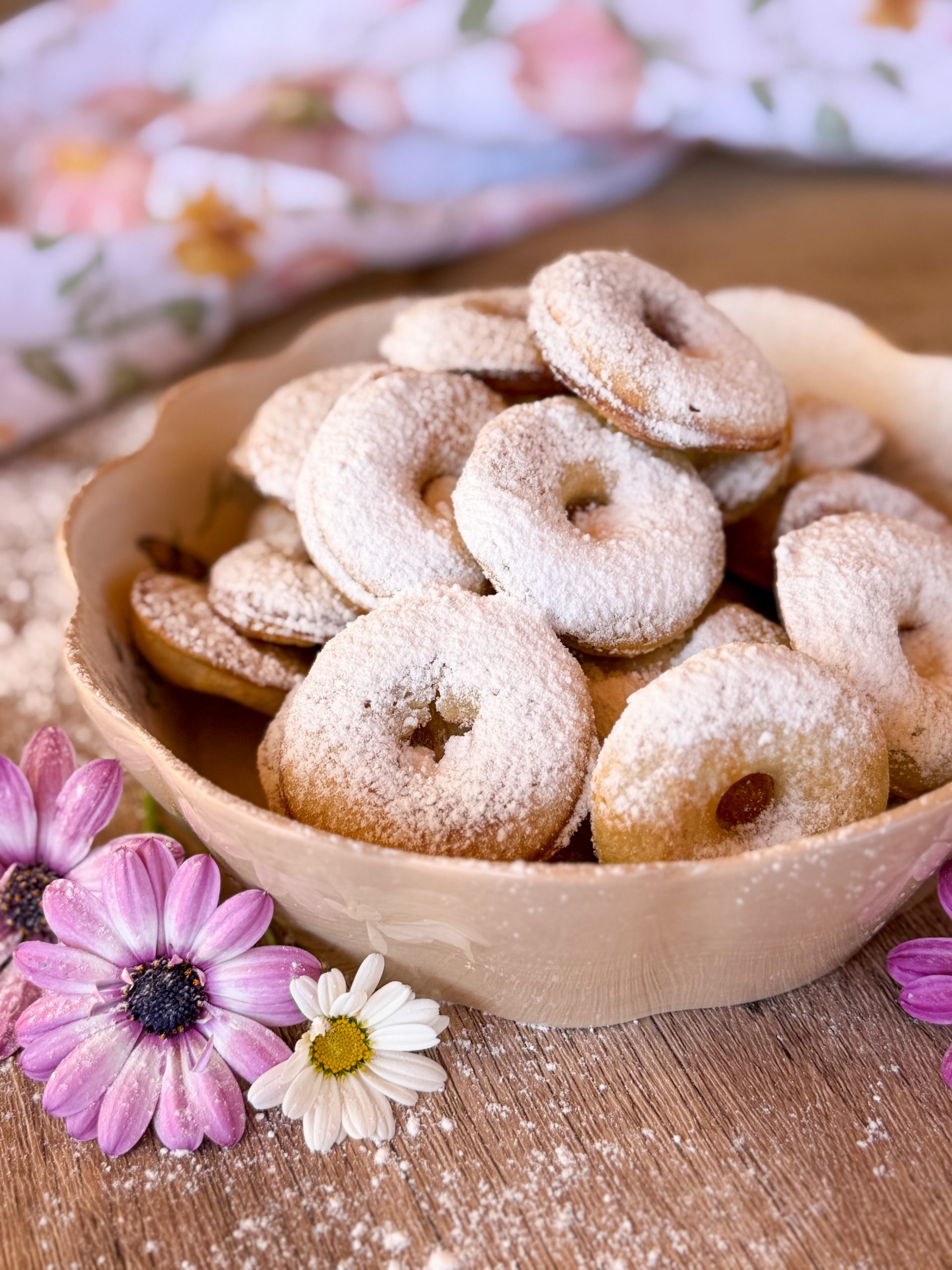 Varios donetes de cheesecake sobre superficie clara con decoración floral. Merienda saludable y fácil de preparar con donut maker.