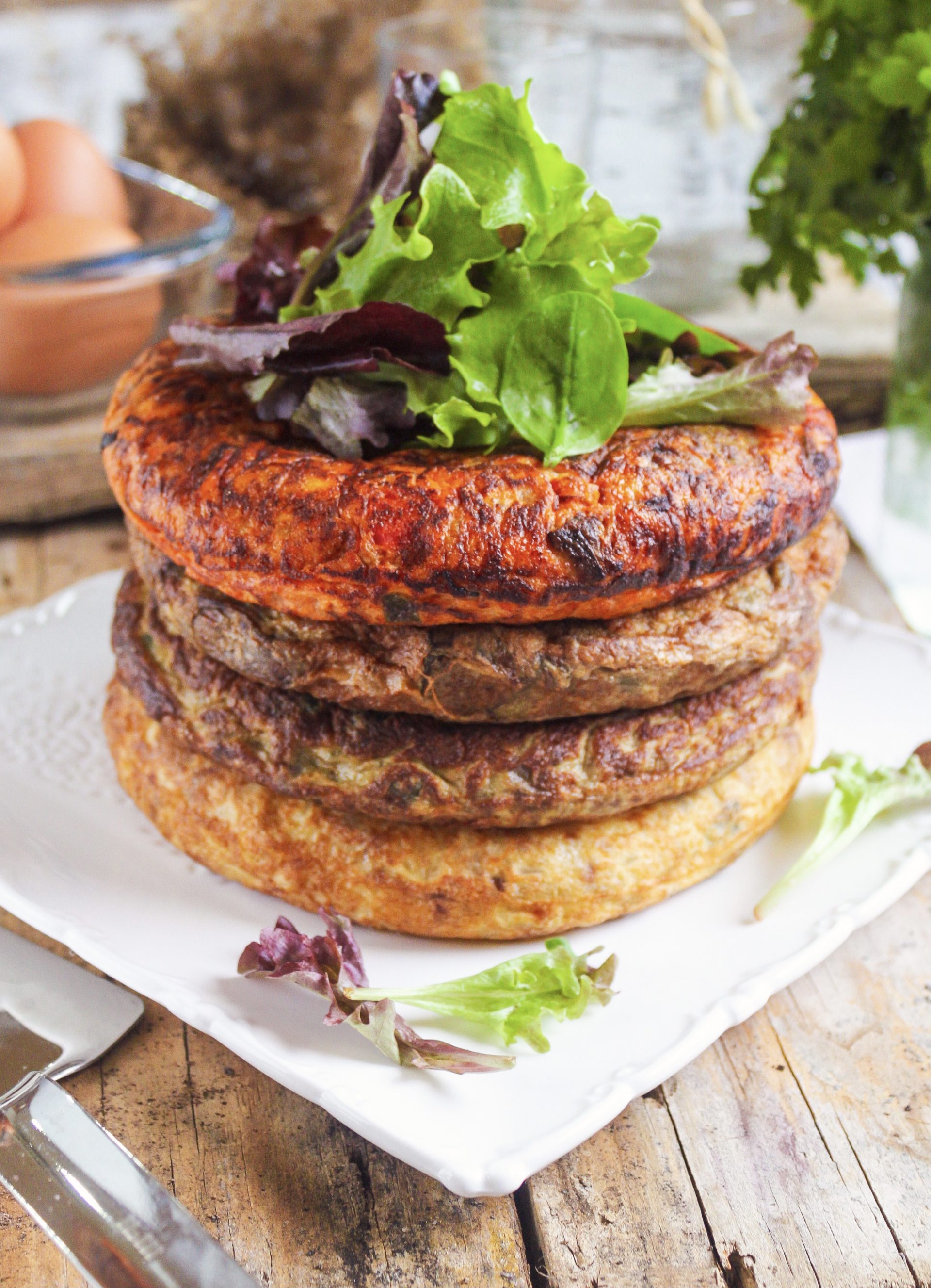 Vista lateral de un pastel de cuatro capas de tortillas individuales (patata, calabacín, alcachofa y pisto) servido en un plato de cerámica blanca sobre una mesa de madera rústica con luz natural y una rama de romero al lado.