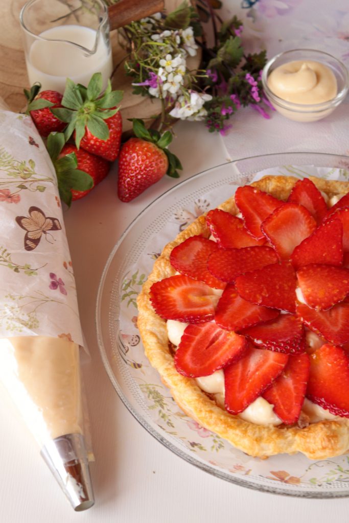 Tarta rectangular de hojaldre crujiente cubierta con una capa generosa de crema pastelera y filas ordenadas de fresas frescas brillantes, dispuesta sobre un papel de horno en una cocina rústica.