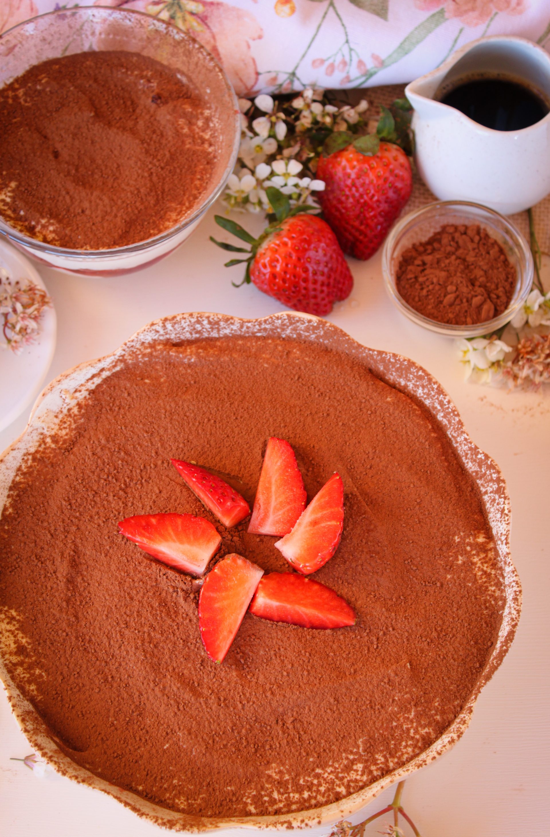 Primer plano de un tiramisú de fresas casero en un cuenco de cerámica blanca. Se aprecian las capas de bizcocho empapado en café, crema de mascarpone y un corazón de fresas naturales cocinadas. Una cuchara de madera muestra la textura cremosa del postre sobre una servilleta de flores sobre una mesa de madera.