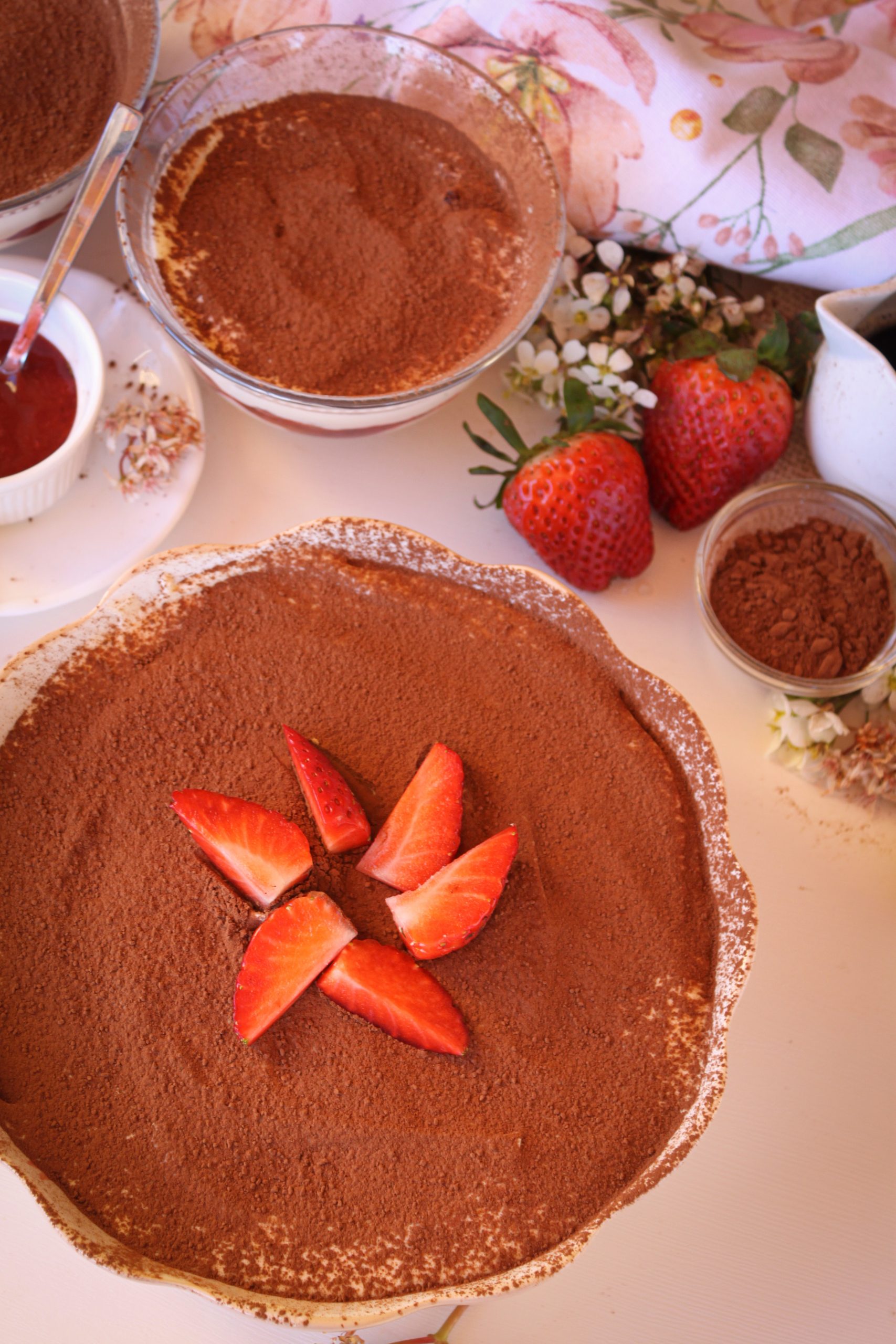 Tiramisú de fresas decorado con cacao en polvo y fresas frescas cortadas. La composición incluye un cuenco rústico, flores de manzanilla blancas y fresas enteras alrededor sobre una mesa de madera iluminada con luz natural de mañana. Fotografía de gastronomía de estilo "Soulfood".