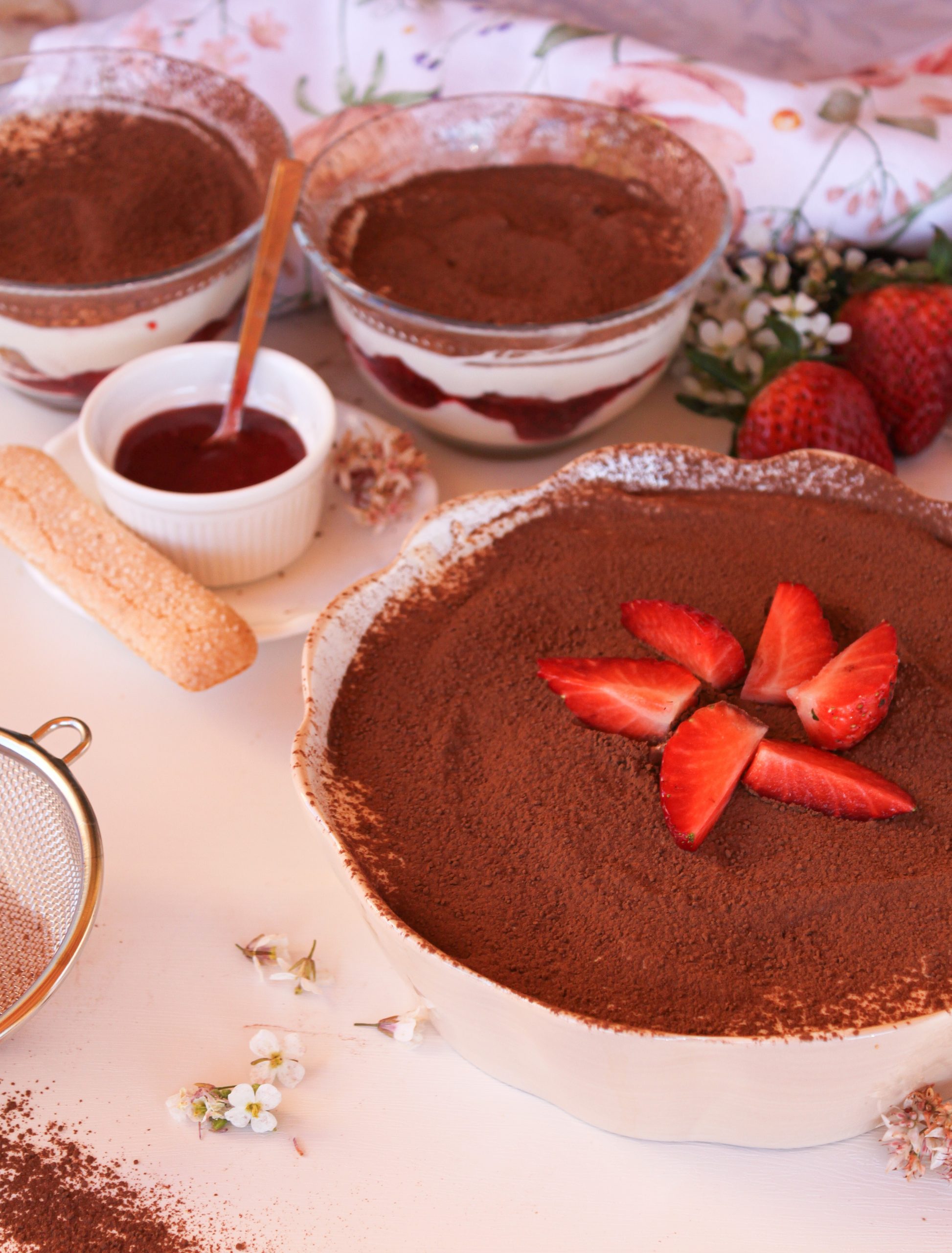 Primer plano de un tiramis&uacute; de fresas casero en un cuenco de cer&aacute;mica blanca. Se aprecian las capas de bizcocho empapado en caf&eacute;, crema de mascarpone y un coraz&oacute;n de fresas naturales cocinadas. Una cuchara de madera muestra la textura cremosa del postre sobre una servilleta de flores sobre una mesa de madera.
