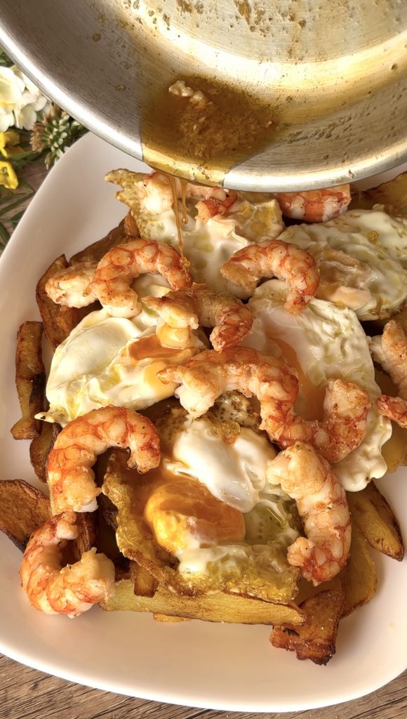 Regando los gambones con el jugo de fre&iacute;rlas en la sart&eacute;n sobre la cama de huevos fritos y patatas.