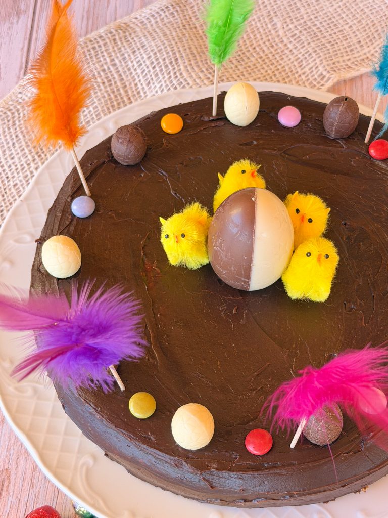 Mona de Pascua catalana artesana con cobertura de ganache de chocolate con leche, decorada con pollitos amarillos, plumas de colores y huevos de chocolate sobre un plato de cerámica blanca.