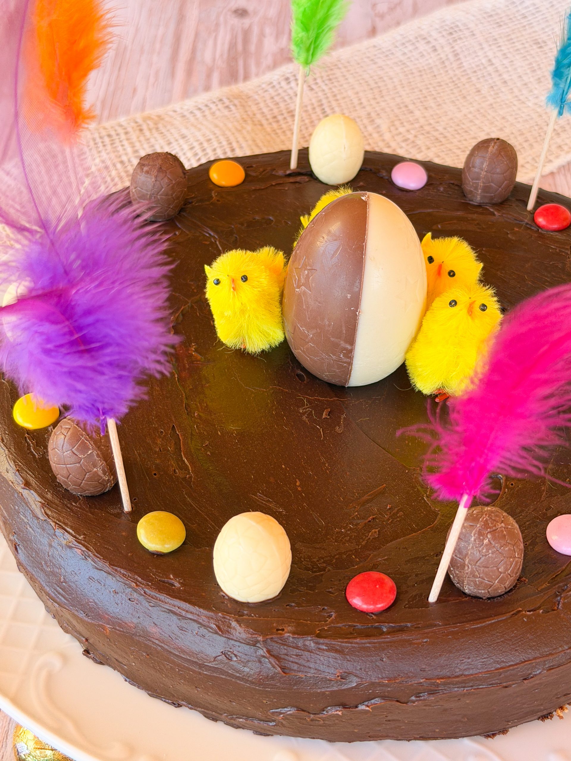 Mona de Pascua catalana artesana con cobertura de ganache de chocolate con leche, decorada con pollitos amarillos, plumas de colores y huevos de chocolate sobre un plato de cerámica blanca.