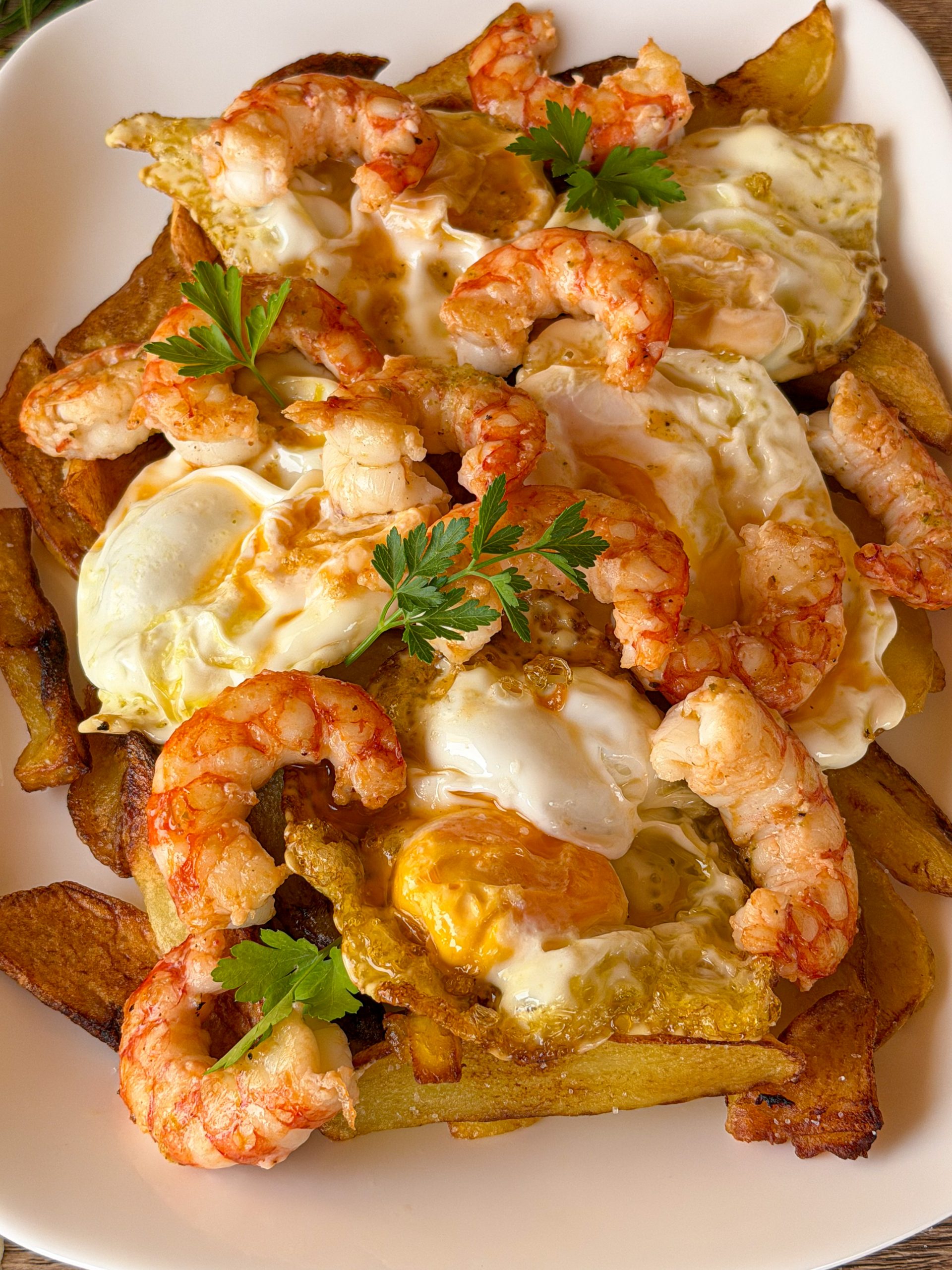 Primer plano de una bandeja de huevos rotos con patatas fritas y gambones pelados, coronada con yemas líquidas y puntilla crujiente, presentada junto a unas ramas de perejil fresco en una mesa de madera rústica