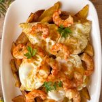 Primer plano de una bandeja de huevos rotos con patatas fritas y gambones pelados, coronada con yemas líquidas y puntilla crujiente, presentada junto a unas ramas de perejil fresco en una mesa de madera rústica