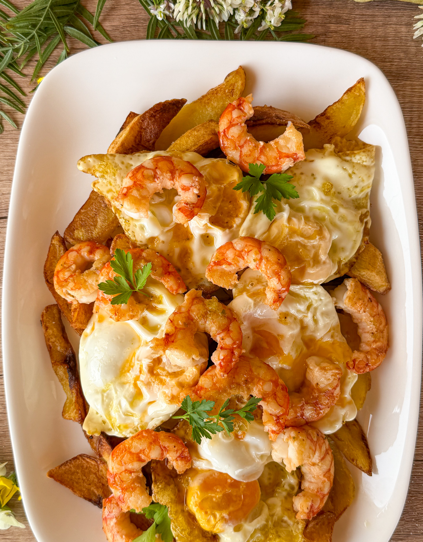 Primer plano de una bandeja de huevos rotos con patatas fritas y gambones pelados, coronada con yemas líquidas y puntilla crujiente, presentada junto a unas ramas de perejil fresco en una mesa de madera rústica