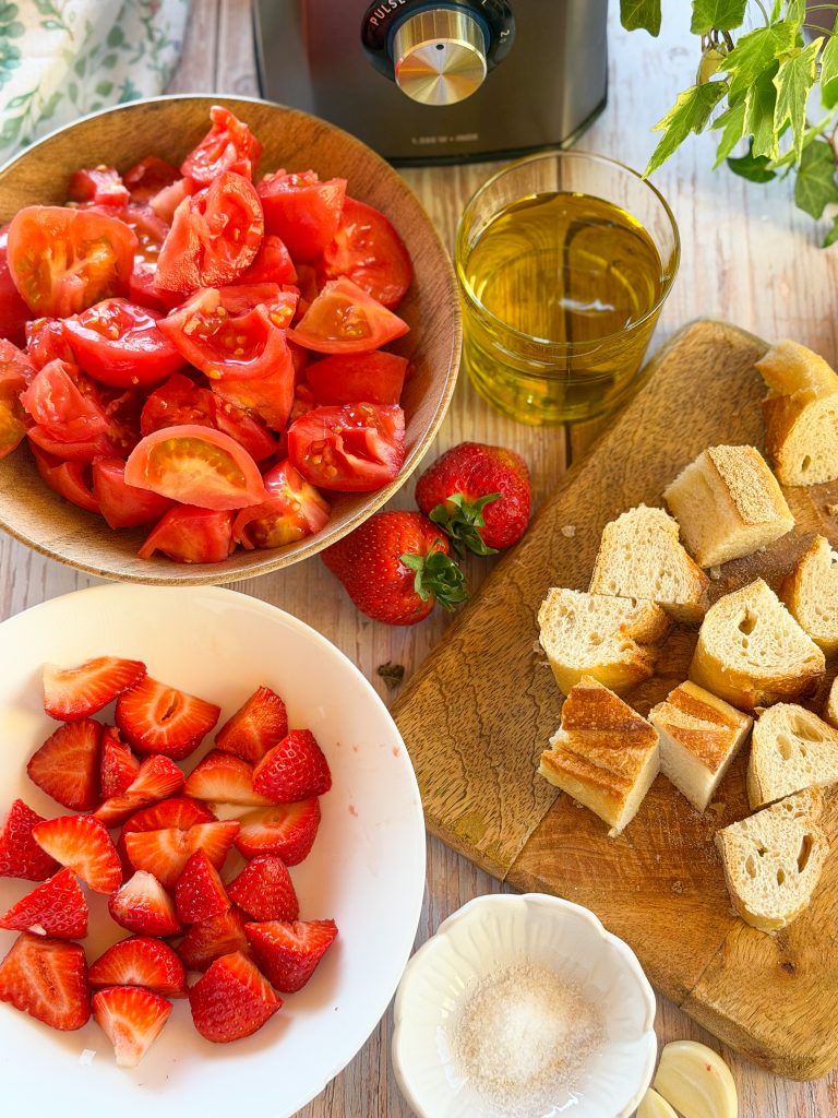 Ingredientes frescos para salmorejo de fresas de temporada: tomates pera maduros, fresas, pan de hogaza, AOVE y ajo sobre mesa r&uacute;stica.