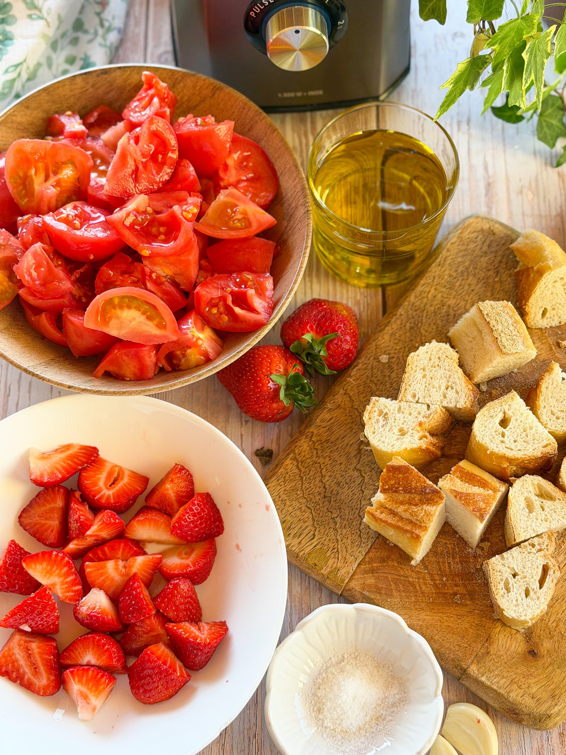 Ingredientes frescos para salmorejo de fresas de temporada: tomates pera maduros, fresas, pan de hogaza, AOVE y ajo sobre mesa rústica.