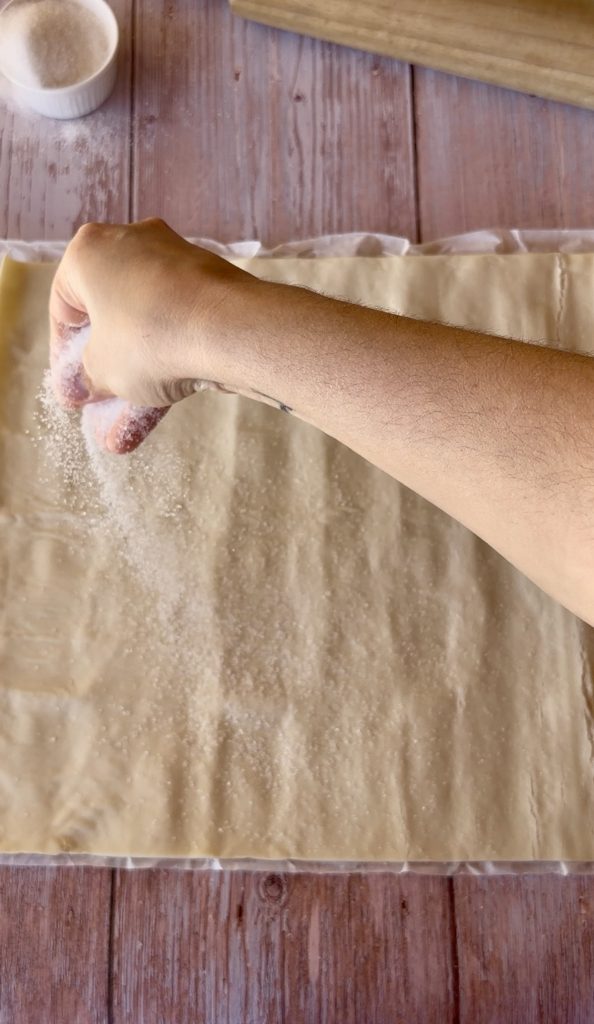 Lámina de hojaldre rectangular extendida sobre una superficie cubierta de azúcar blanco, lista para ser enrollada.