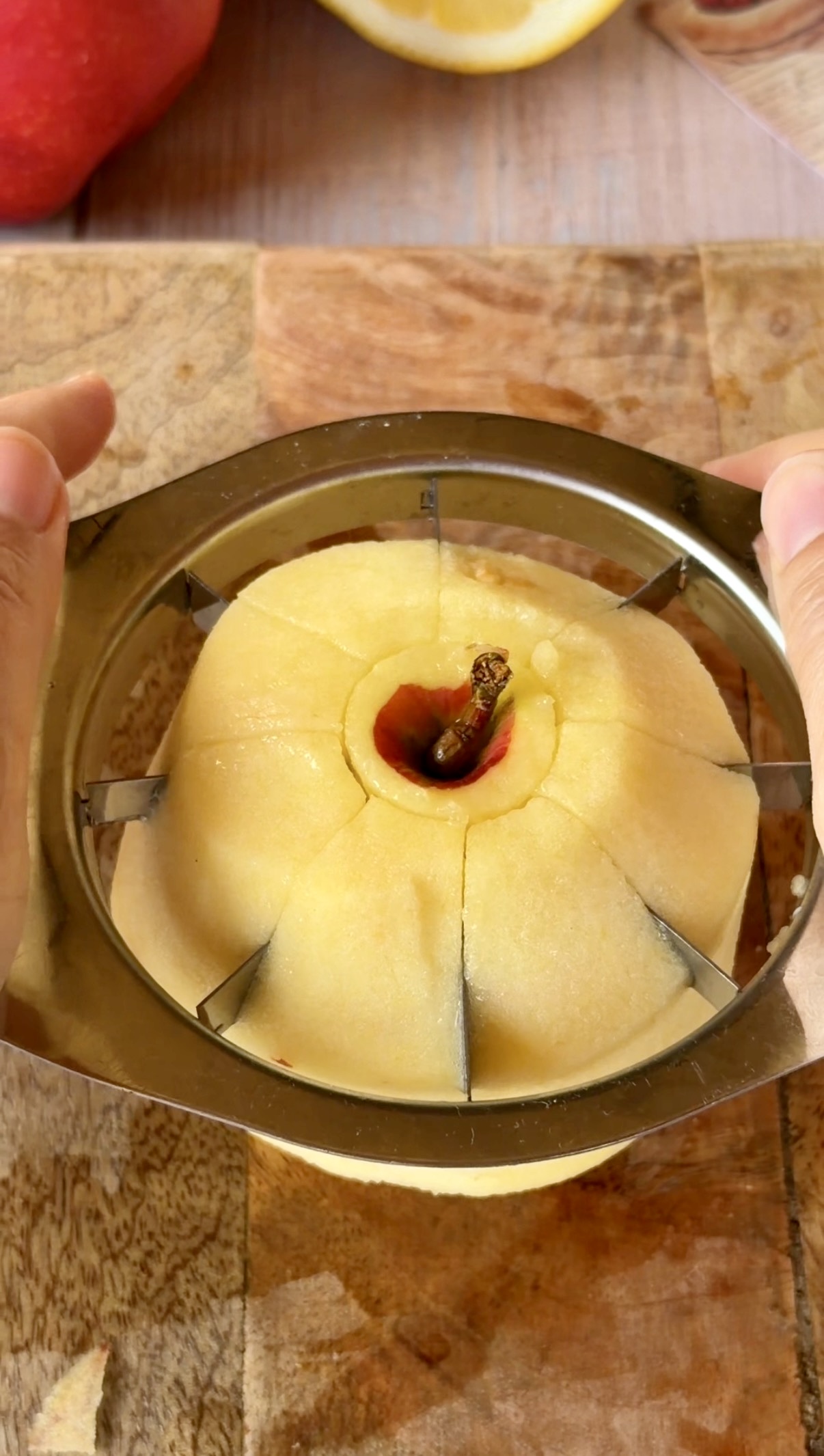 Primer plano de unas manos utilizando un cortador de manzanas metálico sobre una manzana pelada, logrando gajos perfectos y simétricos sobre una tabla de madera.