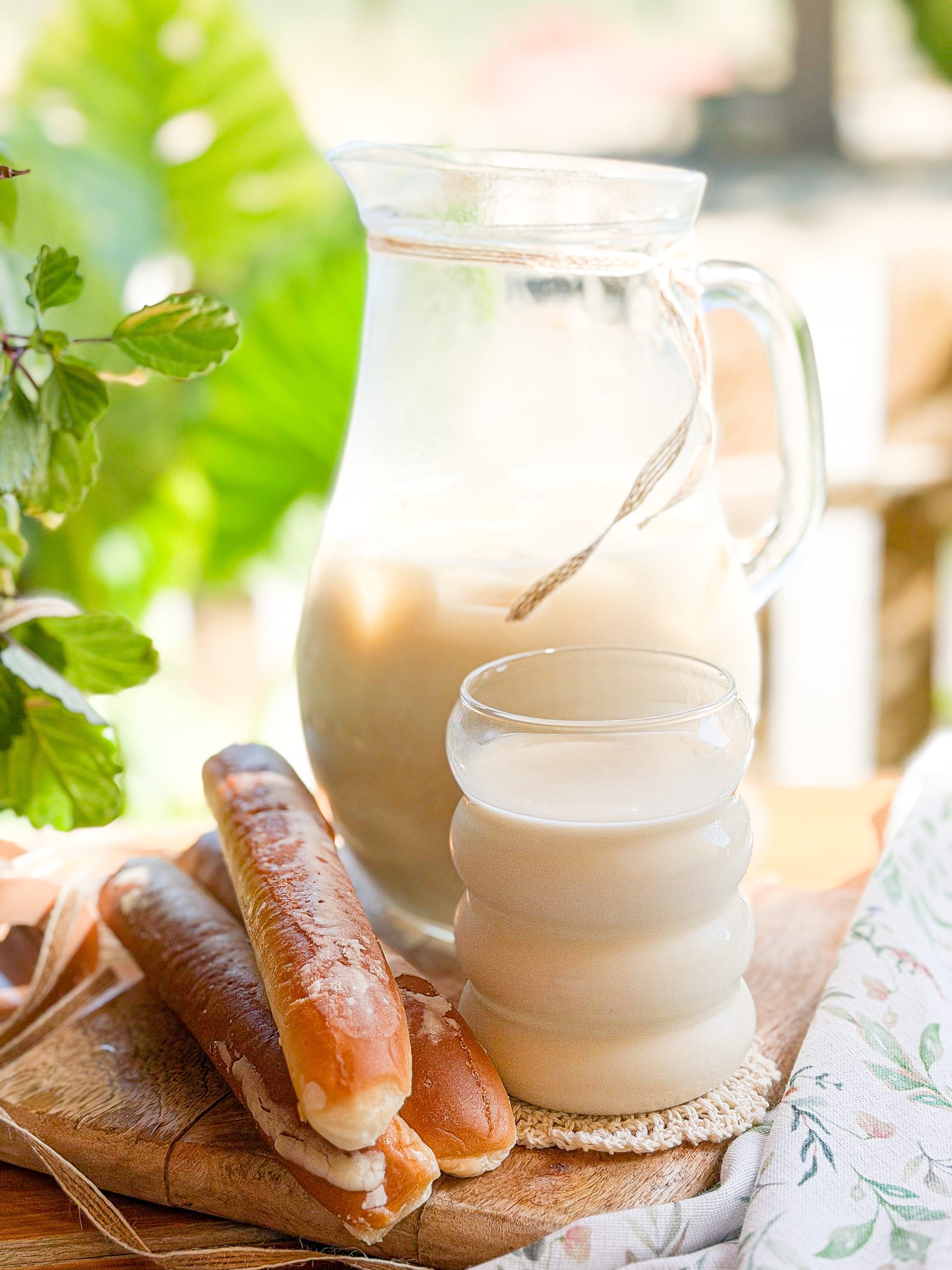 Escena completa de horchata de chufa casera servida en jarra de cristal y vaso, acompañada de fartones sobre una mesa de madera.