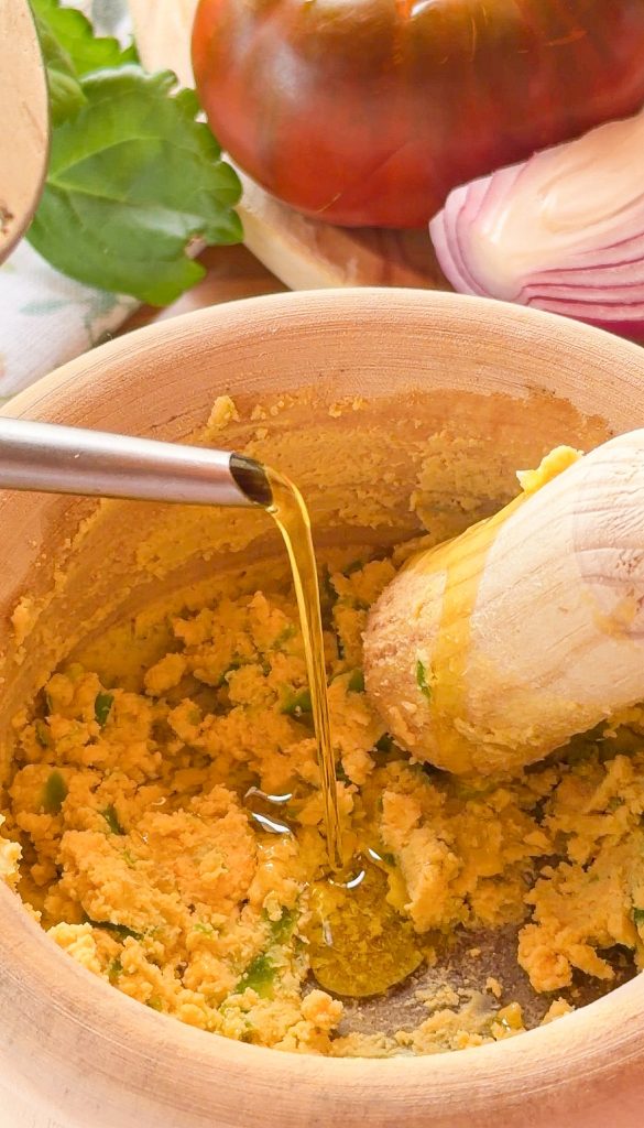 Un chorrito de aceite de oliva virgen extra cae de una aceitera de acero inoxidable sobre el majado de yema, ajo y pimiento verde dentro de un mortero de madera, mientras la mano del mortero está a un lado.