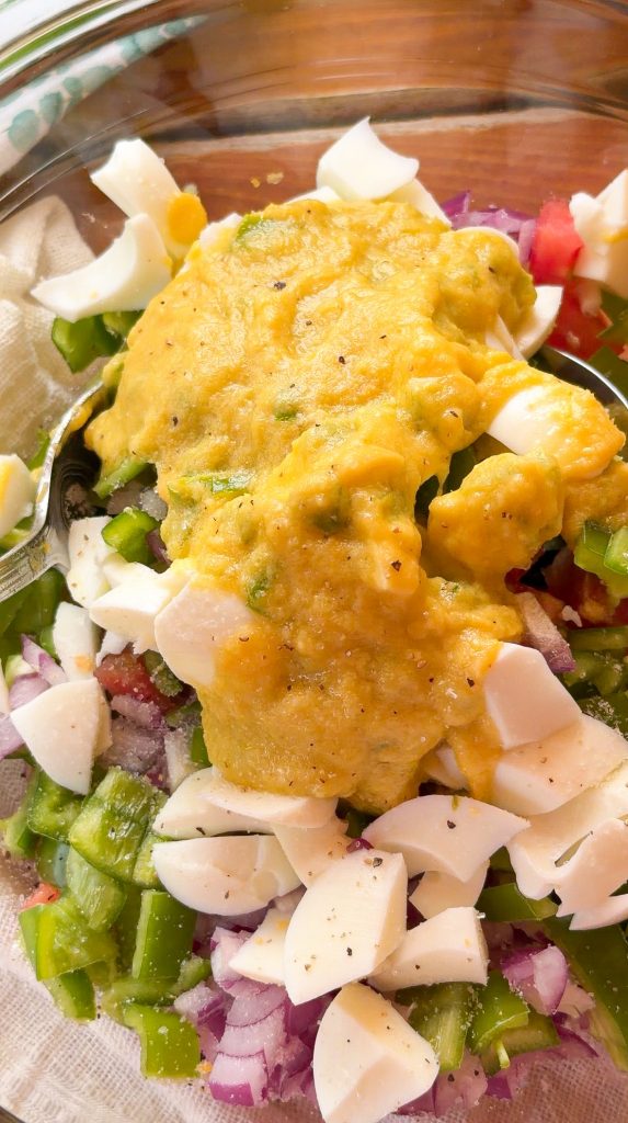Un bol de cristal contiene una base de cebolla roja picada y pimiento verde cortado en dados, coronada con una generosa capa de la emulsión de yema de huevo, aceite y vinagre, lista para ser mezclada.