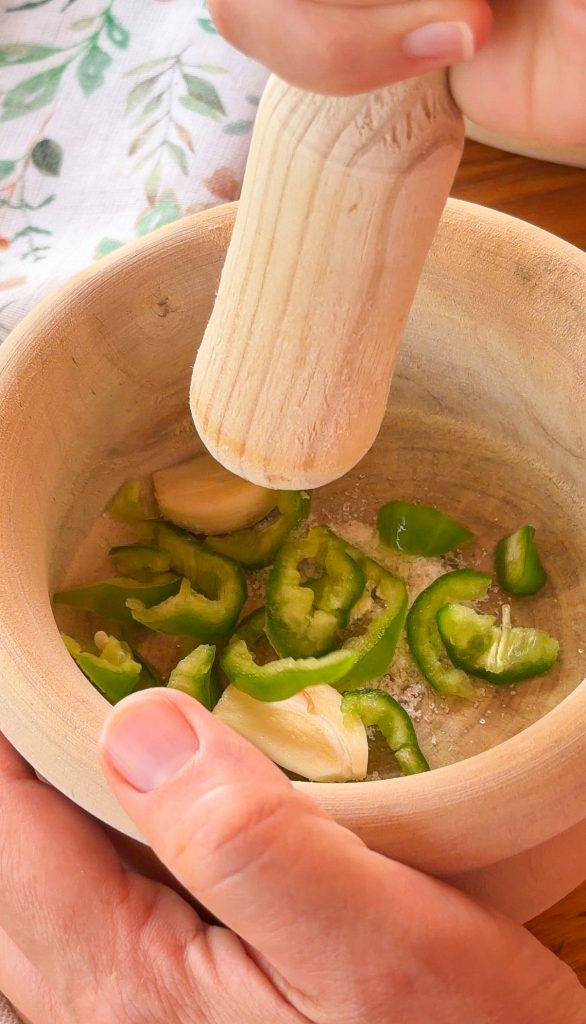 Un mortero de madera contiene trozos de pimiento verde y dientes de ajo pelados, listos para ser machacados con la mano del mortero que se ve arriba.