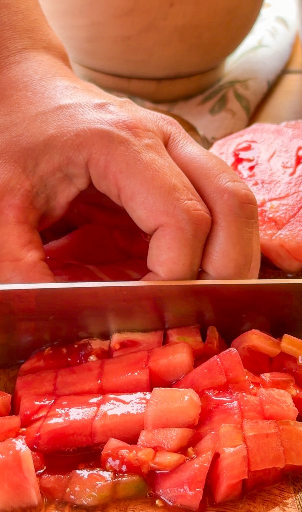 Un cuchillo de cocina corta tomates maduros en daditos pequeños (brunoise) sobre una tabla de madera, con una mano sujetando los tomates y parte de una cebolla al fondo.