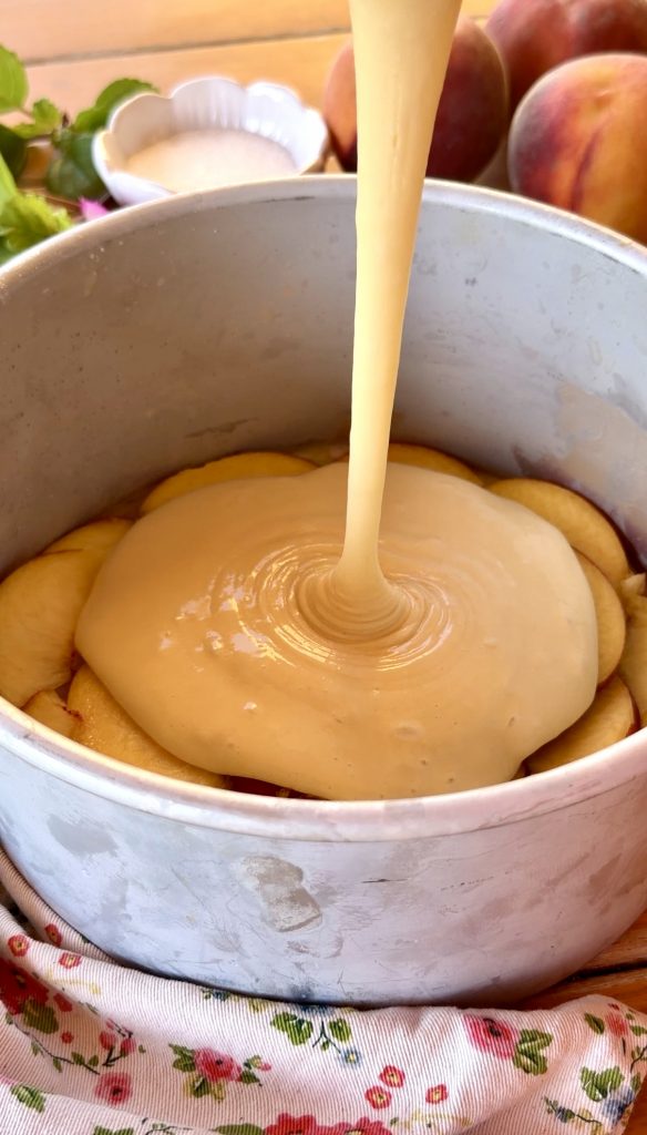 Masa de bizcocho cayendo sobre una base de melocotones caramelizados en un molde redondo antes del horneado.