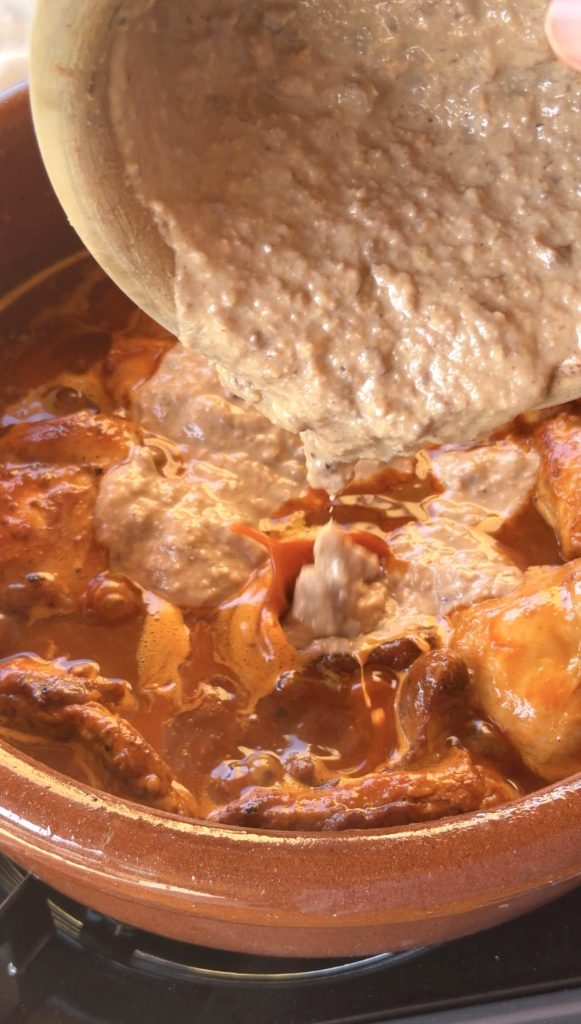 Un cuenco de madera vierte una salsa espesa de color marrón claro, con textura granulada, sobre trozos de conejo dorado que se cocinan a fuego lento en una cazuela de barro llena de una salsa roja vibrante.