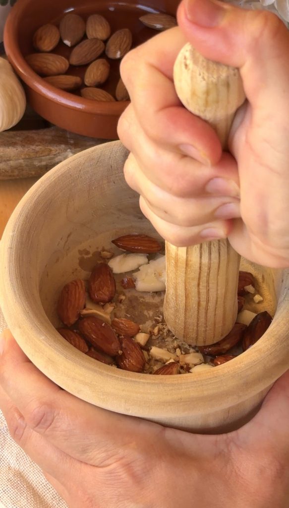 Primer plano cenital de unas manos usando un majo de madera para triturar almendras crudas y otros ingredientes dentro de un mortero de madera. En el fondo, un cuenco de cerámica contiene más almendras.