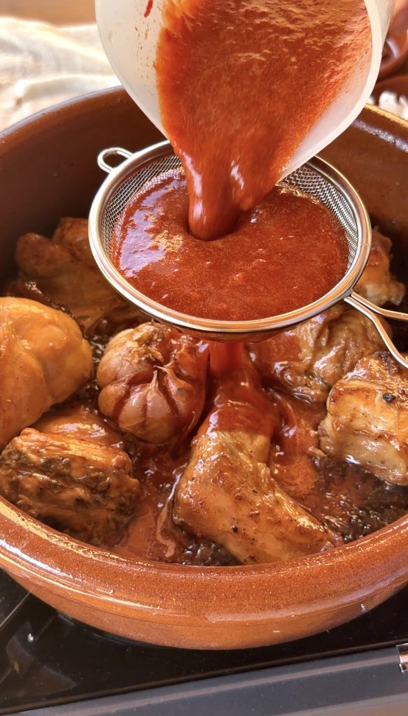 Una mano sostiene un colador de malla fina sobre la cazuela de barro con el conejo dorado y la cabeza de ajos asada. Desde arriba, se vierte una salsa de color rojo intenso a través del colador, que cae sobre la carne.