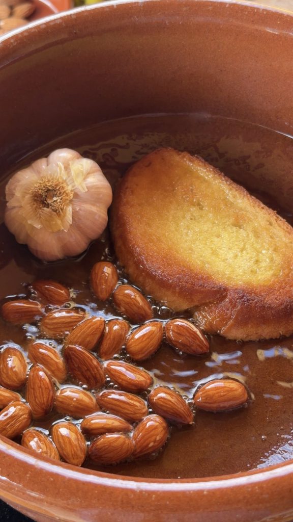 Primer plano cenital dentro de una cazuela de barro con abundante aceite de oliva caliente. En él se fríen una rebanada de pan dorada, una cabeza de ajos entera y numerosas almendras crudas.