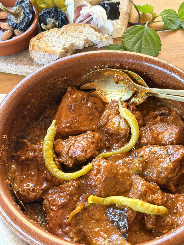 Vista detallada de la cazuela de barro llena con el guiso de conejo terminado. La carne está completamente cubierta por una salsa densa y melosa. Un par de cucharas de servir doradas están hundidas en el guiso. Al fondo, sobre una tabla de madera, hay pan rústico, almendras y ajos secos.