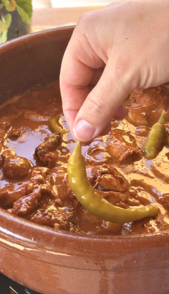 Una mano sujeta por el tallo una guindilla (piparra) en vinagre de color verde amarillento, sumergiéndola parcialmente en el guiso de conejo burbujeante y espeso dentro de la cazuela de barro.