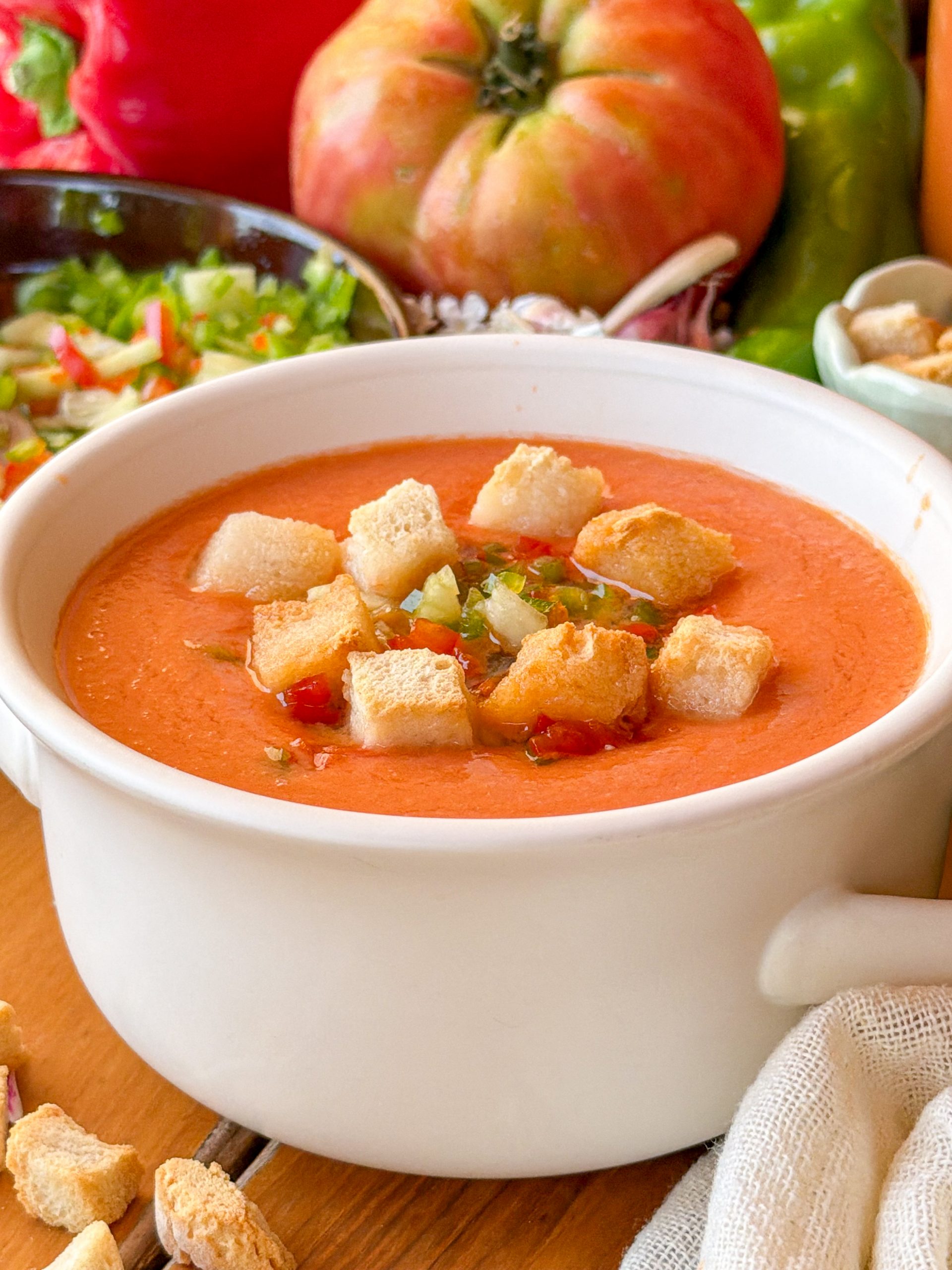 Primer plano de una sopera blanca con gazpacho tradicional decorado con picatostes de pan y verduras finamente picadas, sobre un fondo con tomates frescos y pimientos.