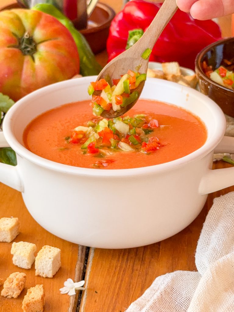 Primer plano de una sopera blanca con gazpacho frío, mientras una mano sostiene una cuchara de madera levantando un poco de guarnición de verduras picadas sobre el plato.