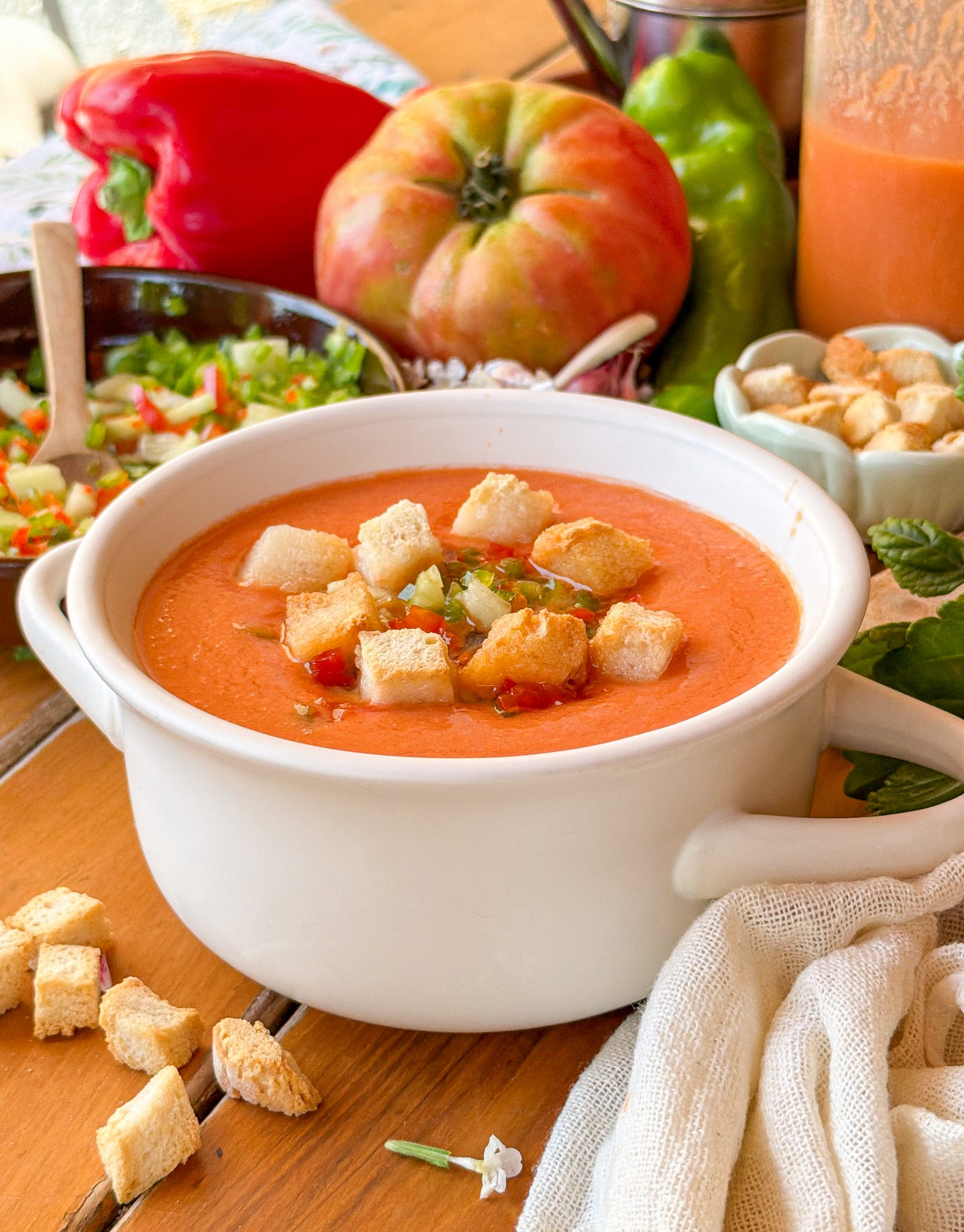 Primer plano de una sopera blanca con gazpacho tradicional decorado con picatostes de pan y verduras finamente picadas, sobre un fondo con tomates frescos y pimientos.