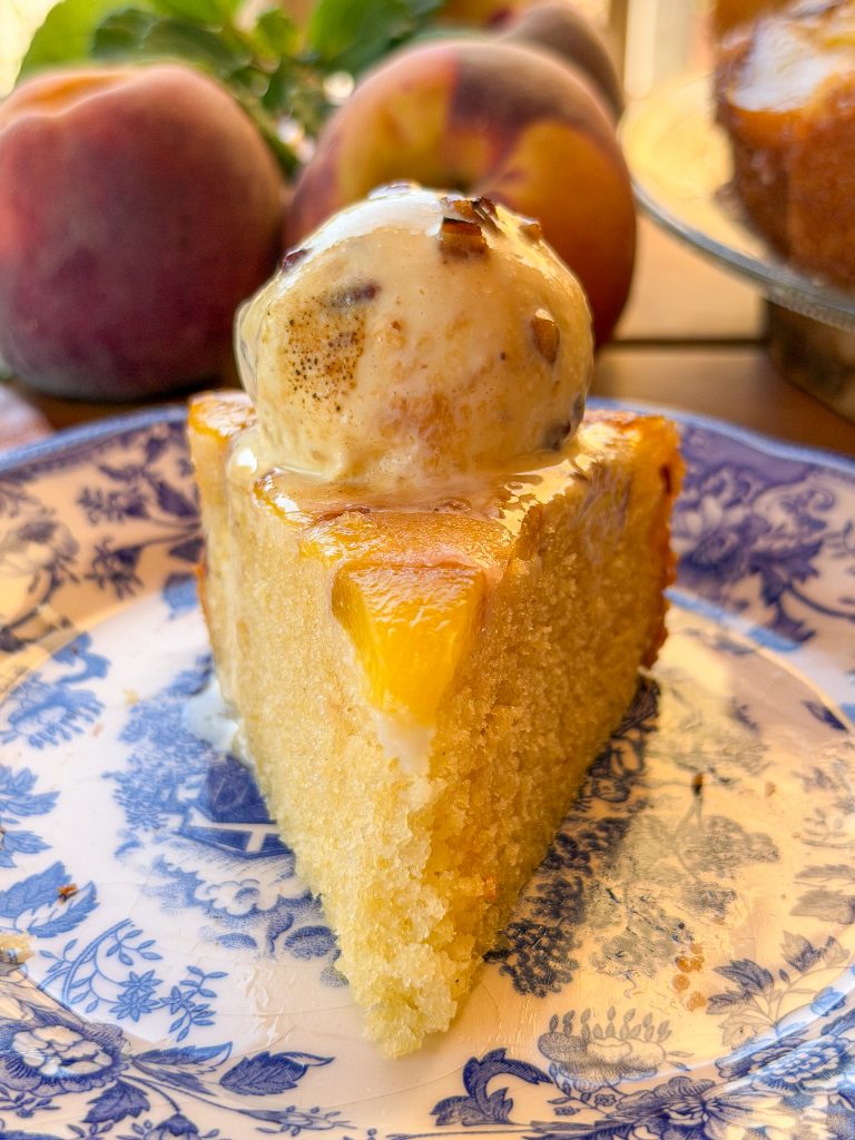 Porción de bizcocho invertido de melocotón caramelizado servida en un plato de cerámica azul, coronada con una bola de helado de vainilla que se derrite lentamente sobre la fruta. Al fondo se ven melocotones frescos y la tarta desenfocada, creando una escena cálida y veraniega.