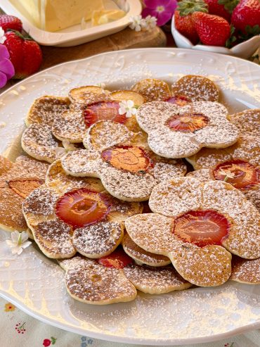 Plato lleno de tortitas caseras con rodajas de fresa en el centro, espolvoreadas con azúcar glass.