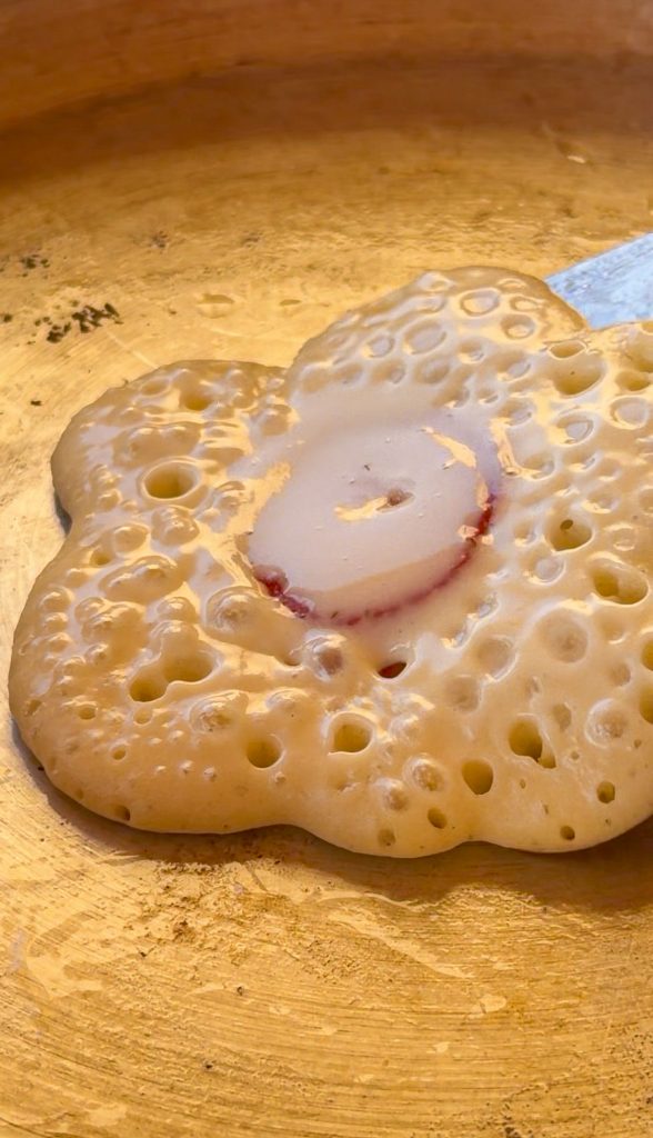 Tortita en forma de flor cocinándose en la sartén, mostrando una rodaja de fresa integrada en el centro y burbujas en la masa.