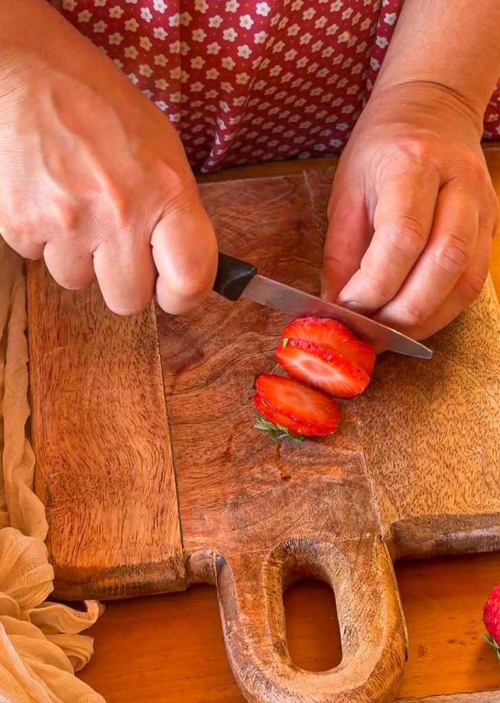 Manos cortando rodajas de fresa fresca sobre una tabla de madera con un cuchillo.