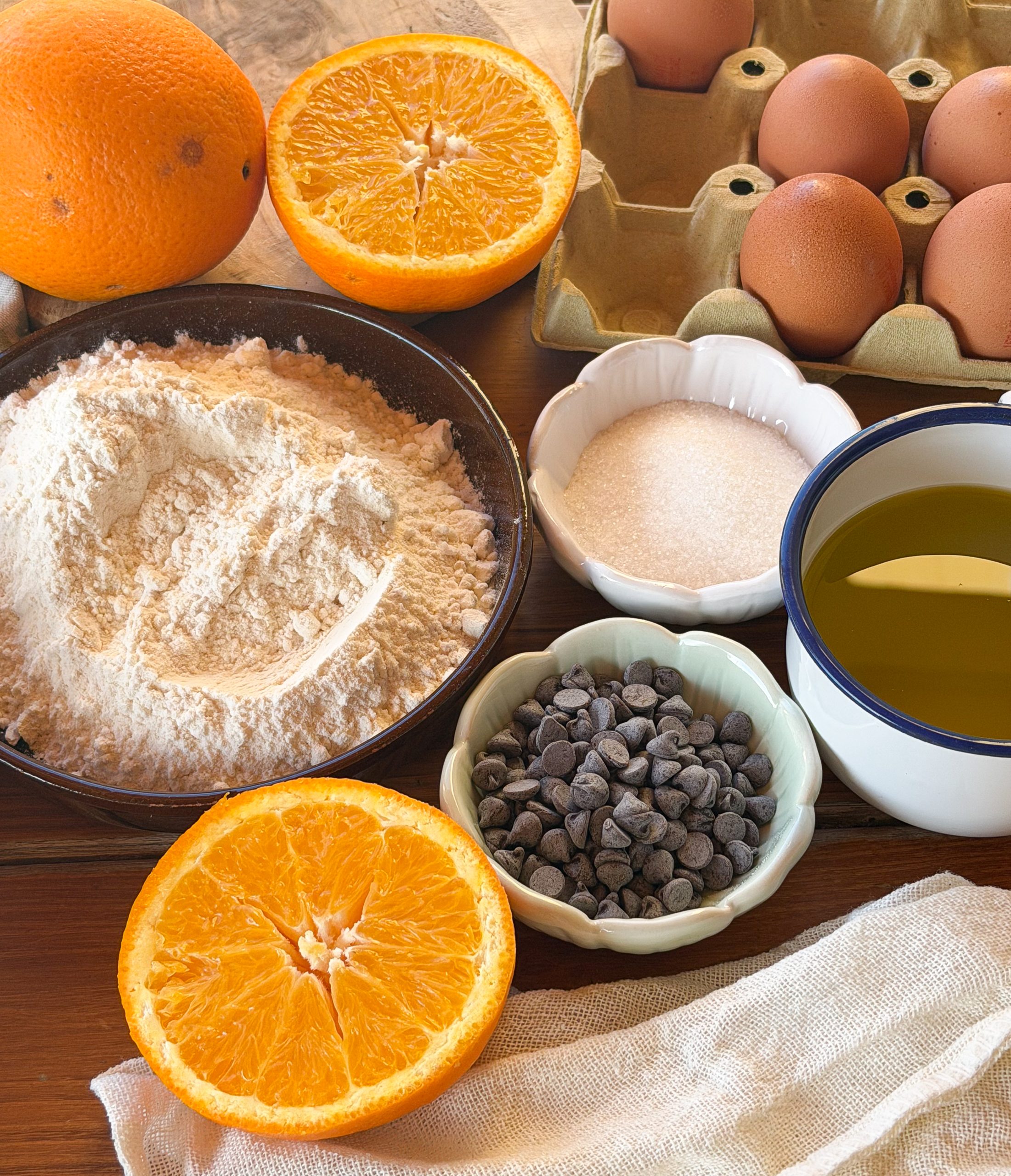 Ingredientes de unas magdalenas caseras de naranja y chocolate con naranja natural, huevos, harina, azúcar, aceite de oliva y pepitas de chocolate listas para preparar la masa.