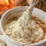 Arroz con leche cremoso con canela molida y una cuchara levantando una porción para mostrar su textura melosa, con velas encendidas al fondo.