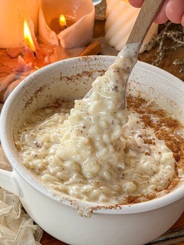 Arroz con leche cremoso con canela molida y una cuchara levantando una porción para mostrar su textura melosa, con velas encendidas al fondo.