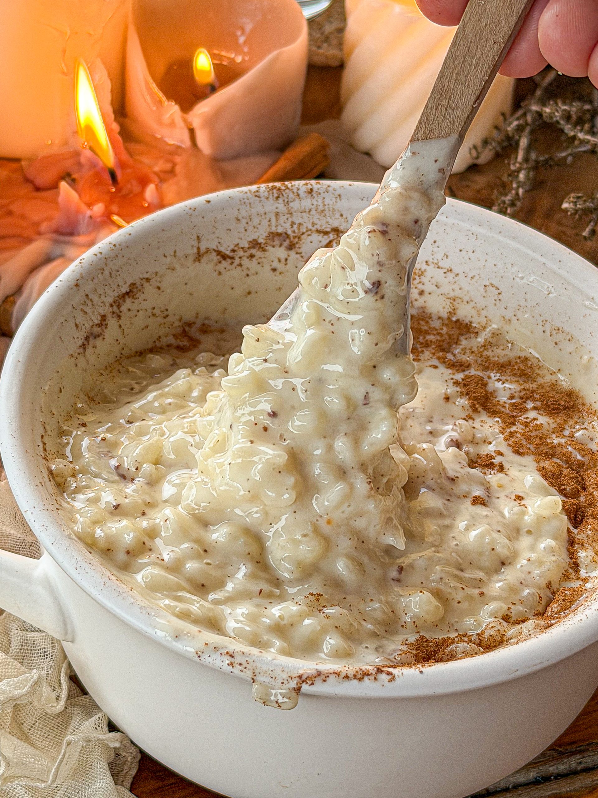 Arroz con leche cremoso con canela molida y una cuchara levantando una porción para mostrar su textura melosa, con velas encendidas al fondo.