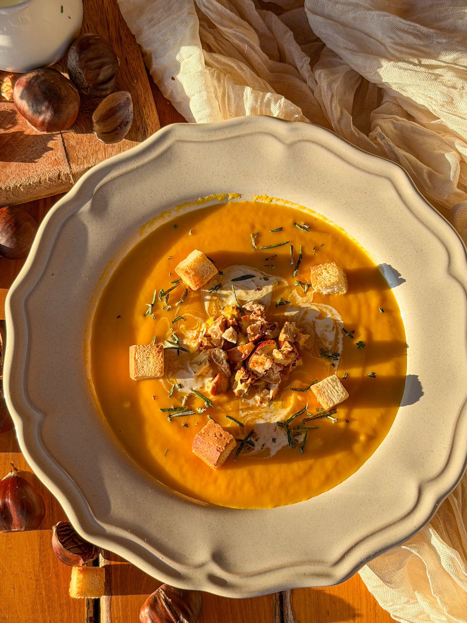 Crema de calabaza asada con castañas picadas, nata, romero fresco y picatostes de pan, servida en un plato rústico.