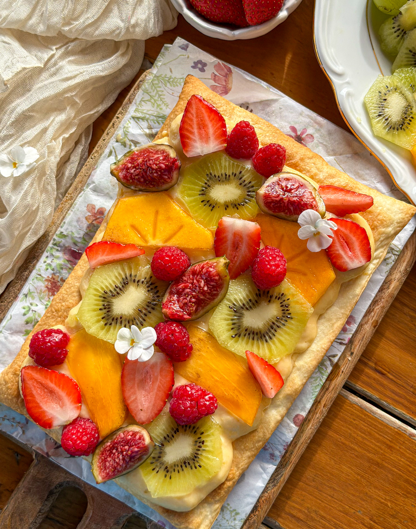 Tarta de frutas frescas con base de hojaldre crujiente y crema pastelera, decorada con kiwi, fresas, frambuesas, caqui e higos.