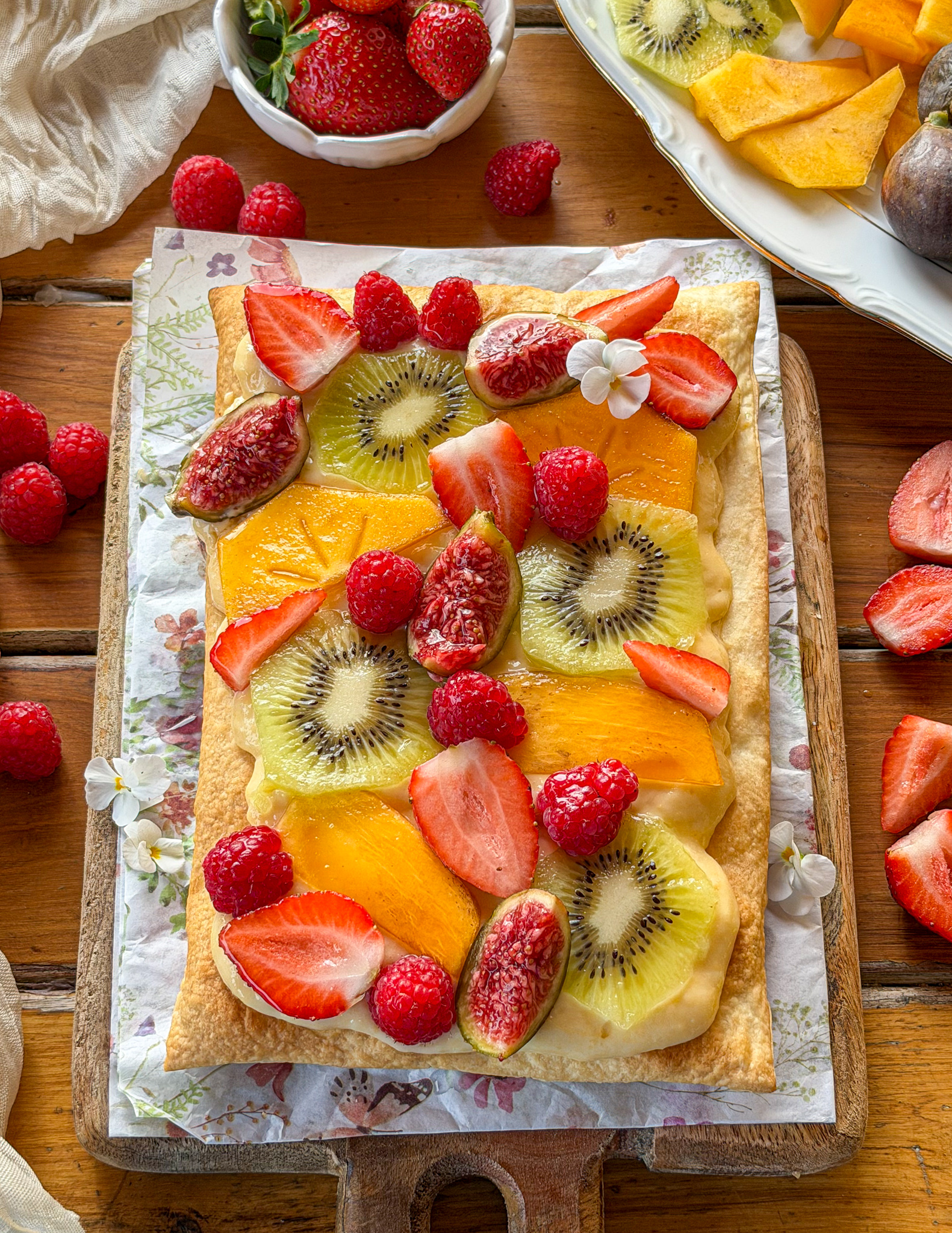 Tarta de frutas frescas con base de hojaldre crujiente y crema pastelera, decorada con kiwi, fresas, frambuesas, caqui e higos.